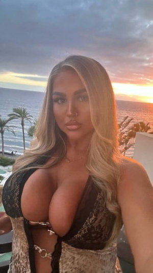 Woman with blonde hair in lace top posing by ocean at sunset.