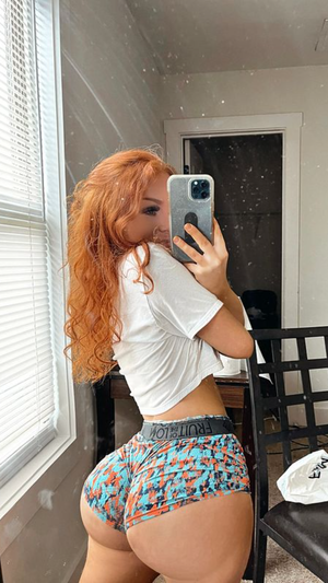Redhead woman in crop top and shorts taking mirror selfie.