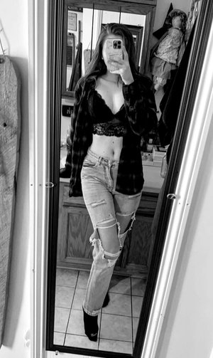 Woman in lace bralette, open plaid shirt, ripped jeans, bathroom mirror selfie.