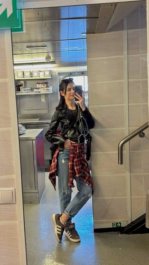 Dark-haired woman in leather jacket taking selfie in kitchen.