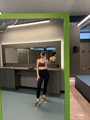 Woman in sports bra and leggings taking mirror selfie in locker room.