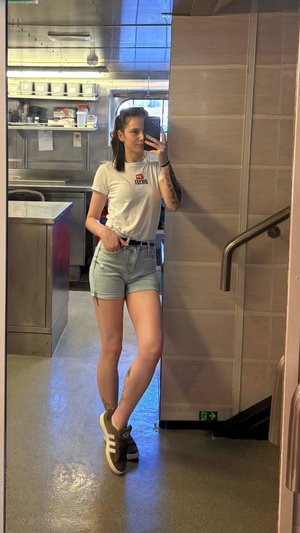 Woman in white t-shirt and denim shorts taking mirror selfie indoors.