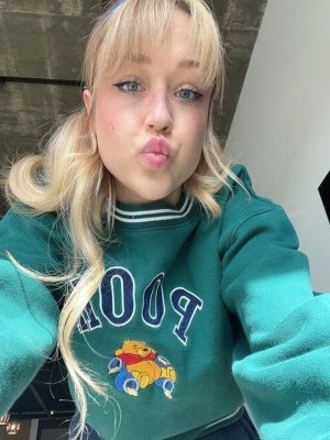 Blonde woman in green sweatshirt with Winnie the Pooh graphic, making a playful face indoors.