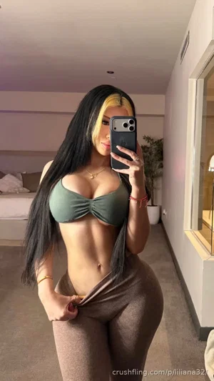 Woman with long black and blonde hair in olive green top and brown pants, taking mirror selfie in