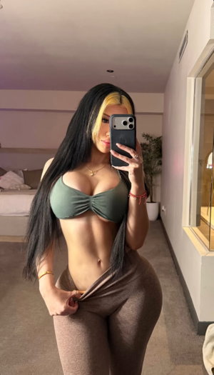 Woman with long black and blonde hair in green top and brown pants, taking mirror selfie in bedroom.