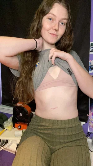 Woman with long brown hair in gray shirt and green pants, posing indoors with hand on neck.