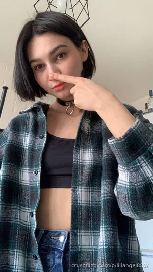 Woman with short dark hair in plaid shirt and black crop top, indoors, pointing at nose.