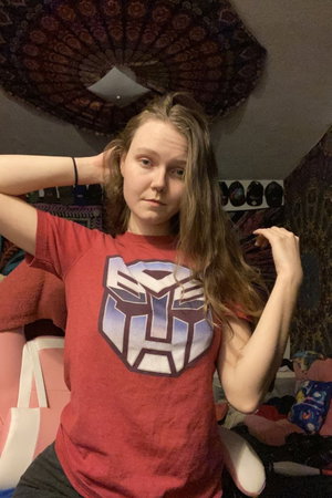 Woman with long hair wearing red t-shirt with Transformers logo, posing indoors.