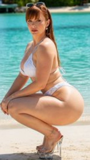 Woman in white bikini squatting on beach near turquoise water.