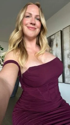 Blonde woman in a maroon off-shoulder dress taking a selfie indoors.