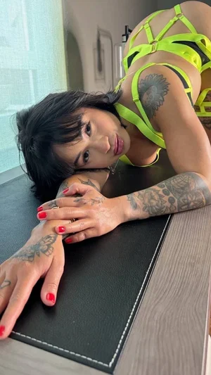 Woman with tattoos in neon green lingerie posing on a black surface indoors.
