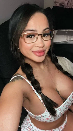 Woman with dark braided hair and glasses wears floral lingerie in a bedroom selfie.