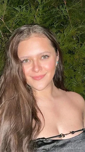 Woman with long brown hair in a strapless top, smiling in front of green foliage.