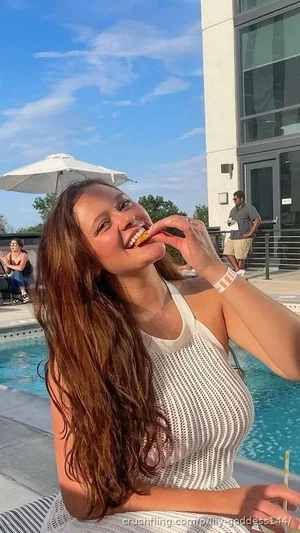 Woman with long brown hair in white mesh top eating by poolside.