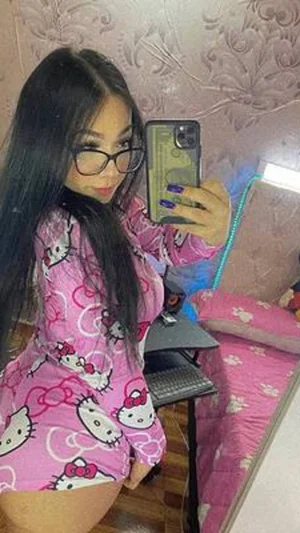 Woman with long black hair in pink Hello Kitty pajamas taking mirror selfie in bedroom.