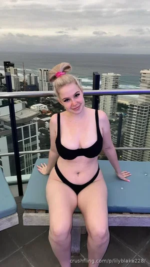 Woman in black bikini posing on balcony with ocean and city view.