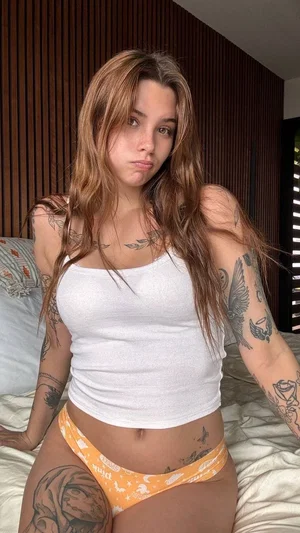 Woman with tattoos in white top and orange underwear sitting on bed indoors.