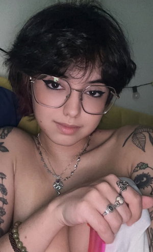 Woman with short black hair, glasses, topless with tattoos, bedroom.