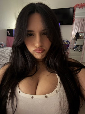 Woman with long dark hair in a white top, posing in a bedroom.