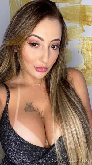 Woman with long brown hair in a black sparkly top, chest tattoo, indoor setting.
