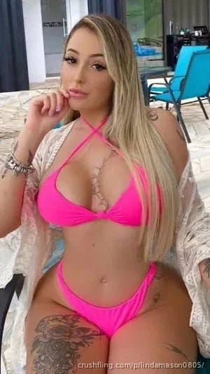 Woman with long blonde hair in pink bikini and white cover-up, sitting outdoors with tattoos