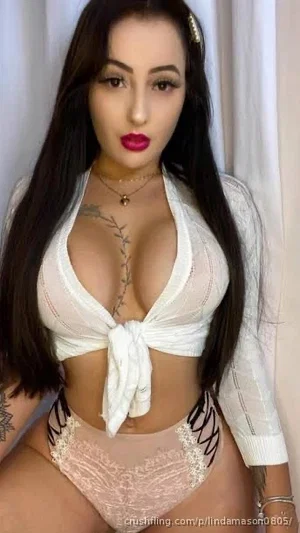 Woman with long dark hair in white lace top and pink lace underwear, standing indoors.