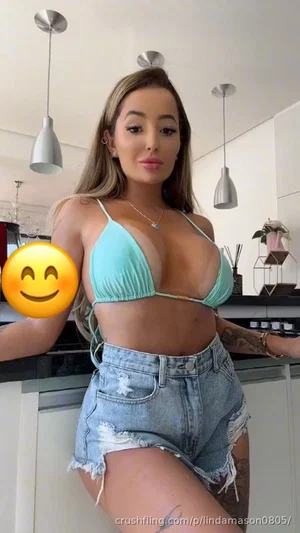 Woman in teal bikini top and denim shorts posing in a modern kitchen.