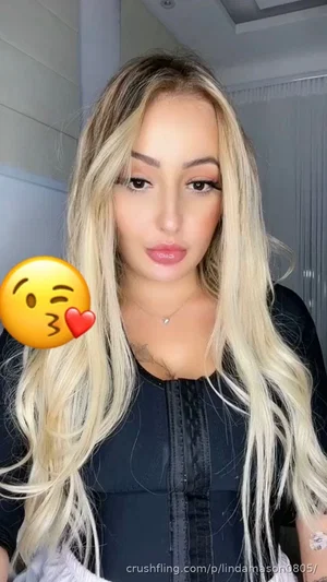 Woman with long blonde hair wearing black top, indoors, selfie with emoji overlay.