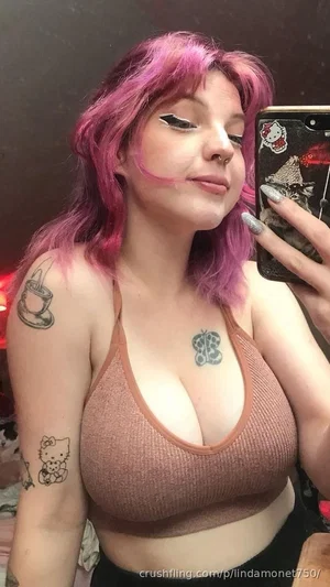 Woman with pink hair and tattoos wears a brown top, taking a mirror selfie indoors.