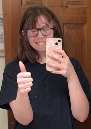 Woman with glasses and brown hair in black shirt giving thumbs up in mirror selfie.