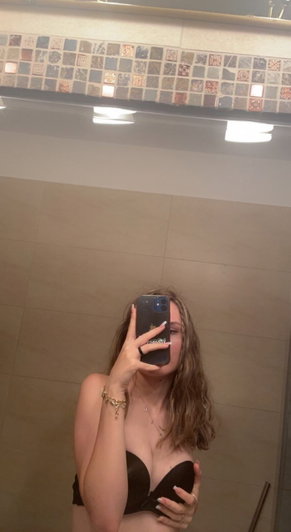 Woman in black bra taking mirror selfie in tiled bathroom.
