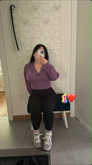 Woman in purple top and black leggings sits on stool, taking mirror selfie in indoor setting.