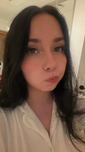 Woman with long dark hair in a white blouse, taking a mirror selfie indoors.