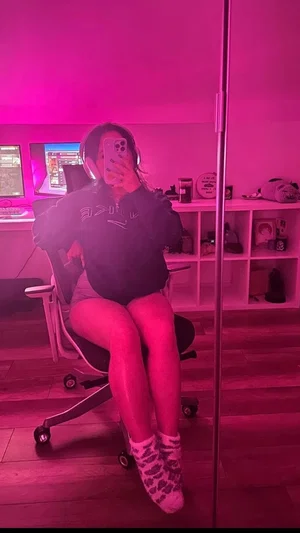Woman in black hoodie and shorts sits in gaming chair, taking mirror selfie in pink-lit room.