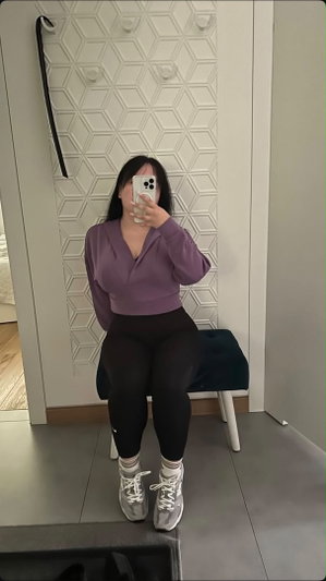 Woman in purple top and black leggings sits on stool, taking mirror selfie indoors.