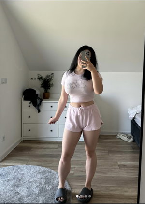 Woman in pink crop top and shorts takes mirror selfie in bedroom.
