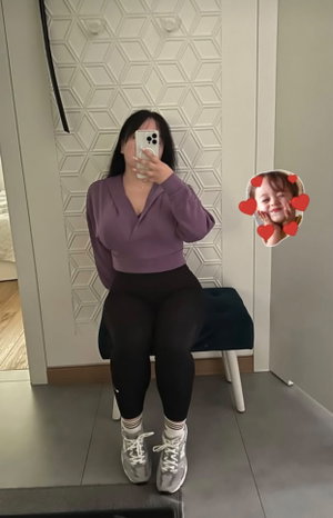 Woman in purple top and black leggings takes mirror selfie in bedroom.