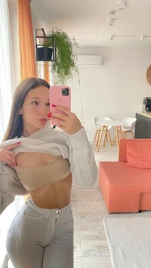 Woman in beige crop top and light pants poses in living room holding pink phone.