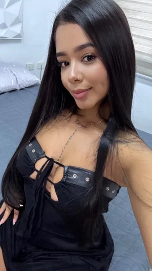 Woman with long black hair in black lace-up top, posing in bedroom.