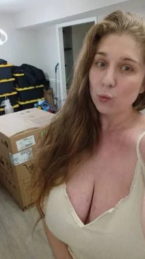 Woman with long hair in a light-colored top, standing indoors near boxes and luggage.