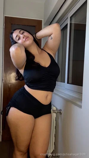 Woman in black crop top and shorts posing indoors near window.