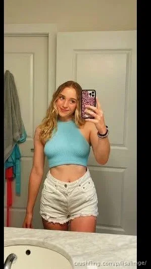 Woman with blonde hair in blue crop top and white shorts taking mirror selfie in bathroom.