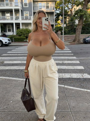 Woman in beige tank top and white pants taking mirror selfie on city street.