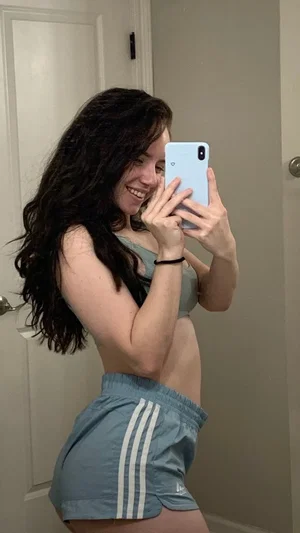 Woman with long dark hair in a gray sports bra and shorts, taking a mirror selfie indoors.