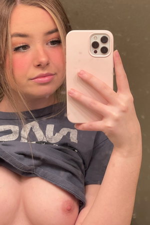 Woman with blonde hair in a mirror selfie, top partially pulled down, showing bare chest.