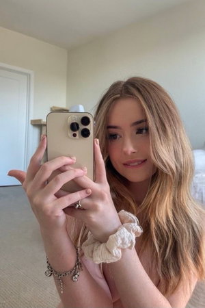 Woman with long blonde hair taking mirror selfie, topless, in indoor setting.