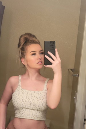 Woman with blonde hair in bun wearing floral crop top, taking mirror selfie indoors.