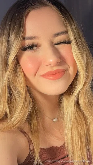 Woman with blonde hair and makeup, wearing a brown lace top, smiling at camera.