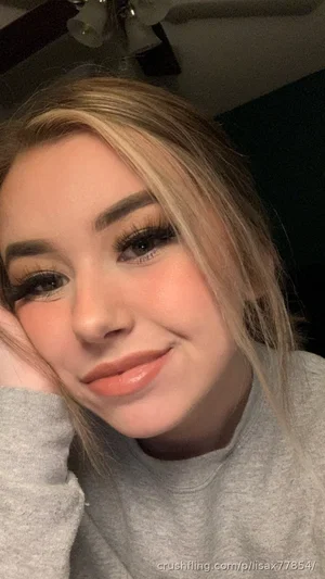 Woman with blonde hair and makeup, wearing a gray sweatshirt, indoors.
