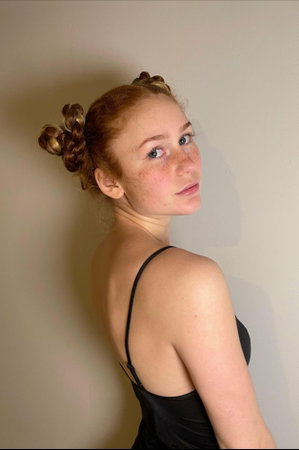 Red-haired woman in black top posing against plain wall.